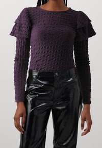 Lila, texturerad långärmad topp med volanger på axlarna, kombinerad med glänsande, svarta byxor med krokodilsmönster.