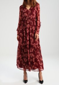 Robe longue bordeaux avec manches longues en voile transparent, motif floral dans des teintes plus claires, taille froncée, ourlet à volants, associée à des talons noirs.