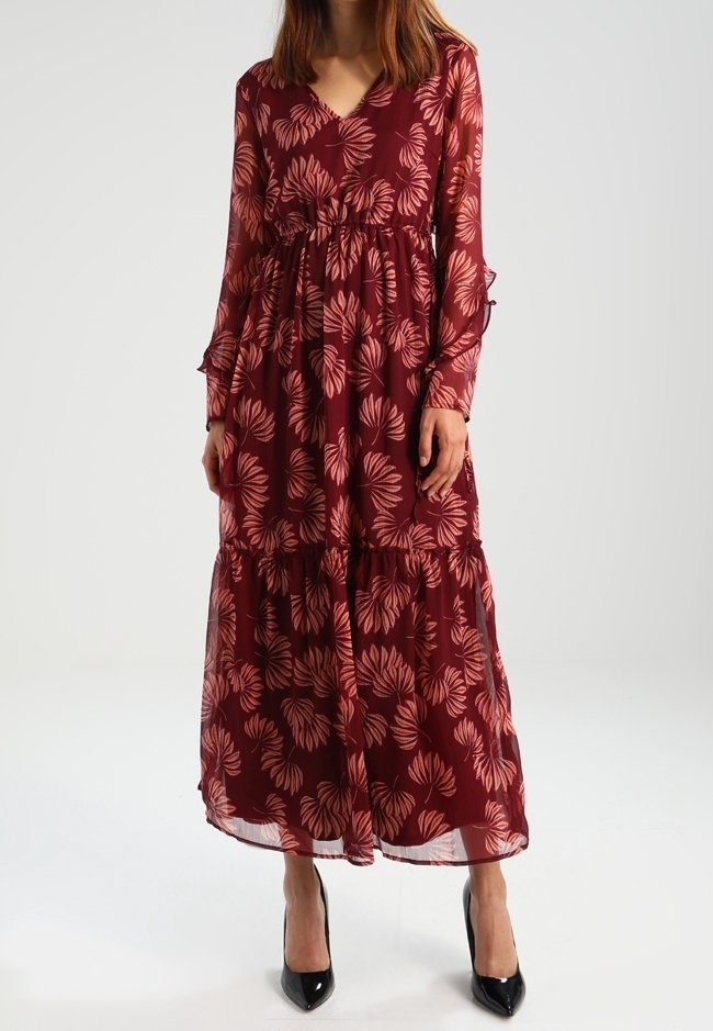 Robe longue bordeaux avec manches longues en voile transparent, motif floral dans des teintes plus claires, taille froncée, ourlet à volants, associée à des talons noirs.