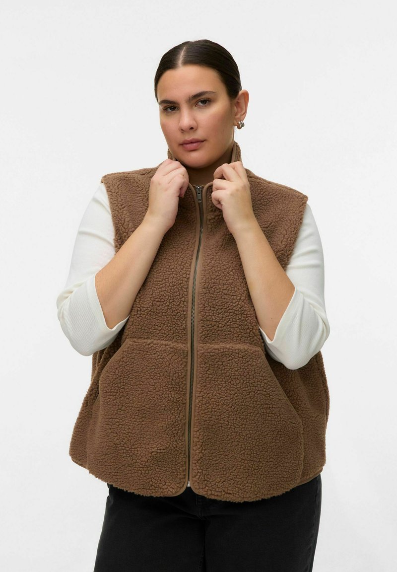 Moteris dėvi rudą vilnos liemenę su užtrauktuku ant baltos ilgomis rankovėmis palaidinės, reguliuoja apykaklę, fone šviesi vientisa sienelė.