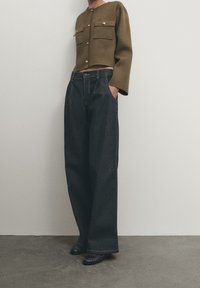 Olive green cropped jacket with gold buttons, paired with dark blue wide-leg denim pants. Textured fabric with side pockets; flat shoes.