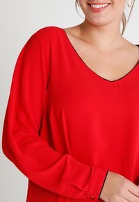 Uśmiechnięta kobieta w jasnoczerwonej bluzie z długim rękawem, z czarnymi wykończeniami na dekolcie i mankietach, z widoczną lekką kolczykiem w kształcie kółka.