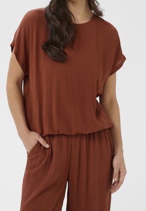Femme portant un haut ample à manches courtes couleur rouille et un pantalon assorti, une main dans la poche, sur un fond uni.