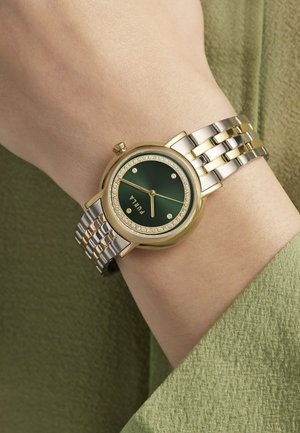 Montre-bracelet en métal doré et argenté avec cadran vert et ornements en cristal portée au poignet sur un tissu vert olive.