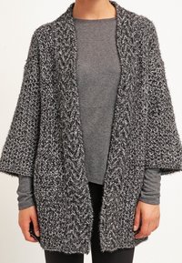 Cardigan aperto nero e bianco lavorato a maglia con maniche larghe, motivo testurizzato e vestibilità ampia. Indossato sopra un top grigio a maniche lunghe.