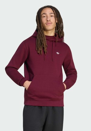 Mand med lange dreadlocks iført en mørkerød Adidas-hoodie med hænderne i frontlommen mod en ensfarvet lys baggrund.