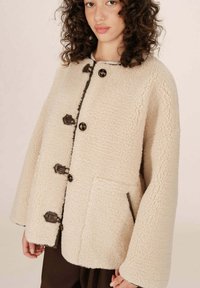 Giacca sherpa color crema, morbida e voluminosa, con una vestibilità rilassata, grandi bottoni marroni e tasche laterali. Presenta un colletto arrotondato e maniche ampie.