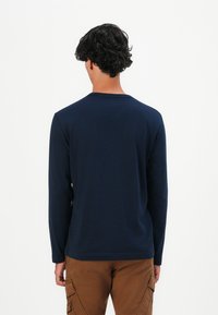 Sötét göndör hajú személy, tengerészkék, hosszú ujjú pólót és barna cargo nadrágot visel, háttal állva egy sima fehér háttér előtt.