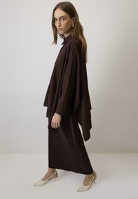 Top marrone a mantella con maniche lunghe e colletto, abbinato a una gonna marrone coordinata. La modella indossa tacchi a punta color crema.