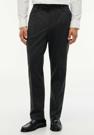 Uomo che indossa pantaloni grigio scuro su misura, mocassini neri e una camicia bianca a maniche lunghe, in piedi con le mani lungo i fianchi.