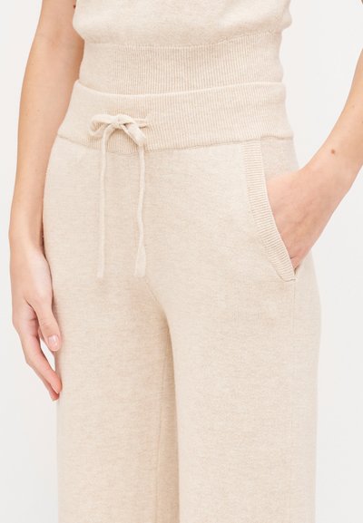 Light beige knit pants with a wide waistband. Features a drawstring tie, side pockets, and a soft, textured fabric.
