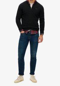 Felpa nera con zip sopra una camicia a quadri, abbinata a jeans blu scuro e sneakers color crema. Caratteristiche notevoli includono polsi e vita a costine.