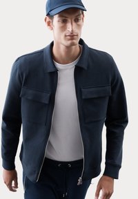 Giacca zip-up blu navy in un tessuto morbido, dotata di due tasche frontali e un colletto, indossata sopra una t-shirt bianca.