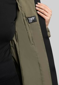 Doudoune verte avec un doublure noire et une étiquette visible sur la couture intérieure. Le tissu semble lisse et légèrement brillant.