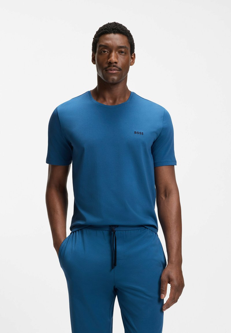 Uomo che indossa una camicia a maniche corte e pantaloni blu coordinati con le mani nelle tasche, in piedi contro uno sfondo bianco uniforme.