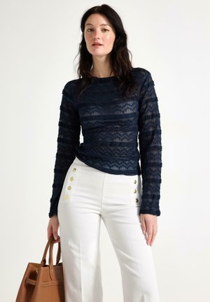 Kvinde iført en marineblå blondebluse med lange ærmer og hvide højtaljede bukser med guldknapper, der holder en brun lædertaske.