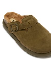 Olijfgroene suède instapklomp met zijklikker en tan kleurig faux bont voering, ontworpen voor comfort en warmte binnenshuis.