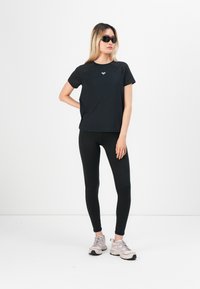 Svart t-skjorte med kort erm og et subtile logo, kombinert med svarte leggings og lyserosa joggesko. Bakgrunnen er ensfarget hvit.