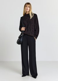 Dunkler grob gestrickter Pullover mit hohem Kragen, kombiniert mit schwarzen Hosen mit weitem Bein. Hält eine kleine schwarze Tasche und trägt spitze schwarze Schuhe.