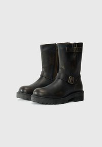 Botas de cuero negras hasta la mitad de la pantorrilla con suelas gruesas, cremalleras laterales y correas decorativas con hebillas en el tobillo y en la parte superior del caño.