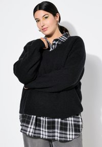 Czarny, oversize sweter dzianinowy z dekoltem w serek, nałożony na czarno-białą koszulę flanelową w kratę. Sweter ma prążkowaną fakturę i rękawy.