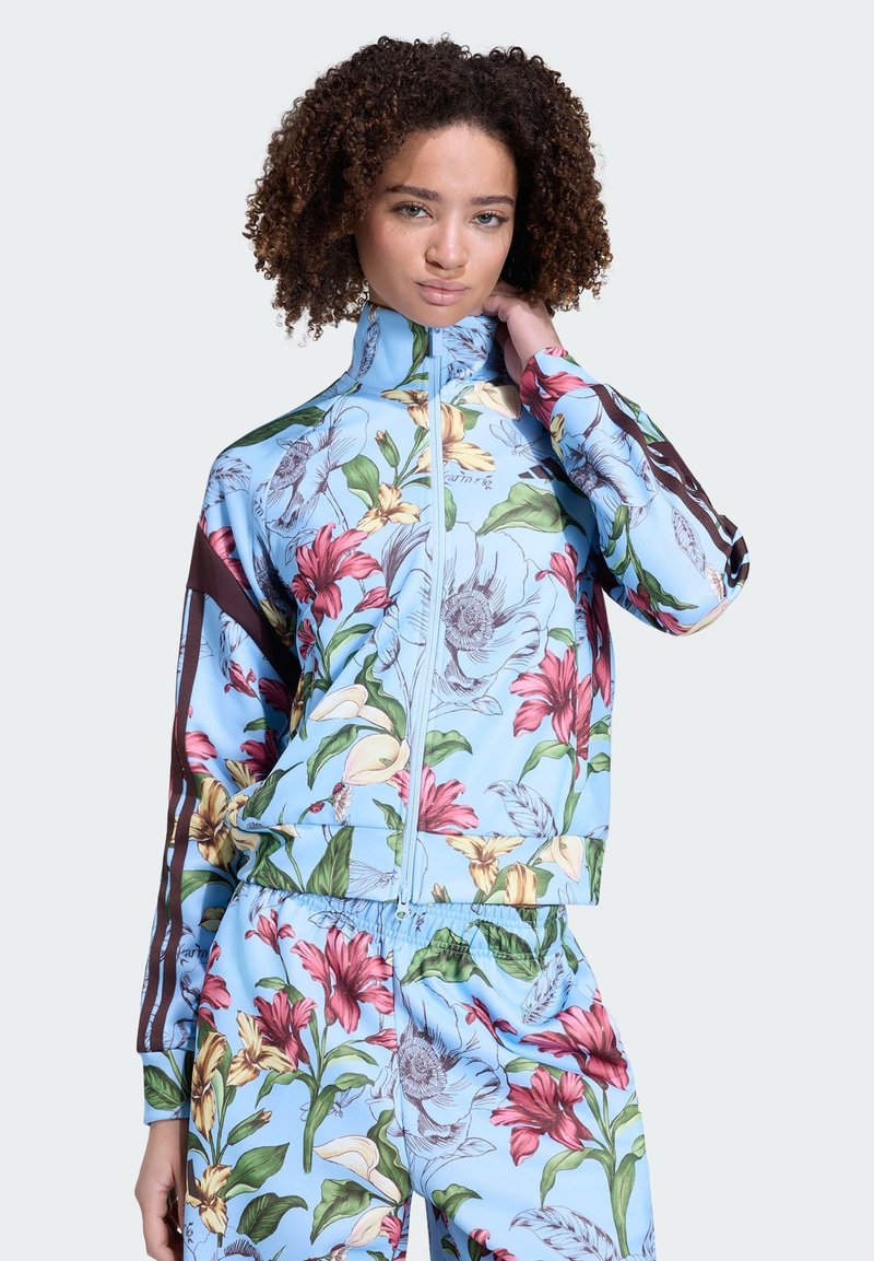 Femme aux cheveux bouclés portant un survêtement bleu clair à fleurs roses, jaunes et vertes, posant avec une main près du cou.