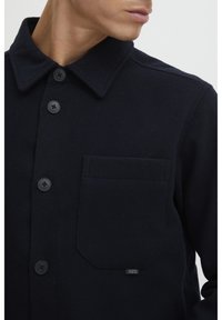 Navy wool blend jacket with a pointed collar, front button closure, and a chest pocket featuring a small label. Soft texture.