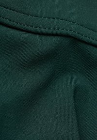 Tmavě zelená látka s hladkým povrchem a jemným žebrováním. Vybavena prošitou lemem s minimálním leskem. Detailní záběr zvýrazňuje vlákna.