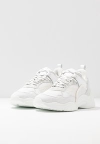 Baskets blanches présentant un mélange de matériaux lisses et texturés, une forme arrondie, un design à lacets et des accents gris clair contrastants.