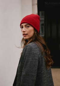 Red ribbed knit beanie on a woman with long brown hair, wearing a textured grey blazer, against a plain background.