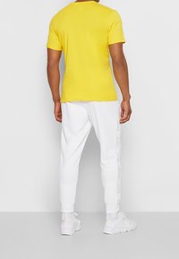Yellow short-sleeve t-shirt, fitted design, paired with white joggers featuring a side pattern. White athletic shoes complete the outfit.