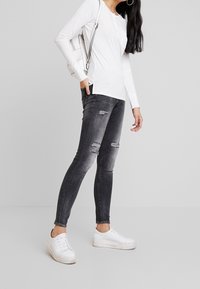 Cienkie dżinsy w ciemnoszarym denimu z podartymi kolanami, w połączeniu z długim, białym topem i białymi trampkami. Prosta torba zawieszona na ramieniu.