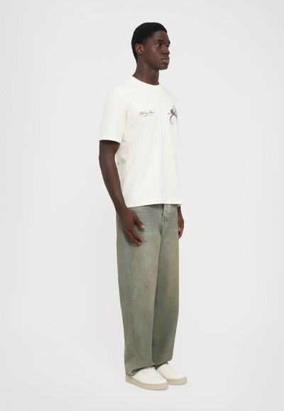 Young man wearing a cream graphic t-shirt, loose faded green jeans, and white sneakers standing against a plain light background.