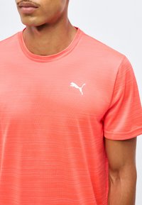 Άνδρας που φοράει ροδακινί ροζ μπλουζάκι Puma με διακριτικό οριζόντιο ριγέ σχέδιο και λευκό λογότυπο Puma στο στήθος, ορατό από τον λαιμό μέχρι το μέσο του κορμού.