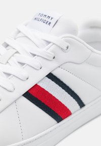 Białe sneakersy ze skórzaną cholewką, z czerwonym i granatowym akcentem w paski. Sznurówki są białe; logo na języku to "TOMMY HILFIGER."