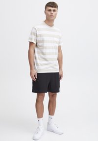 Camiseta de algodón a rayas en beige y blanco con un bolsillo en el pecho, combinada con pantalones cortos texturizados negros y zapatillas blancas.