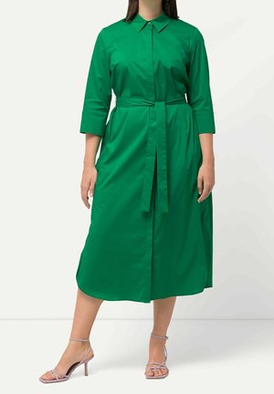 Mujer con un vestido midi verde de manga larga abotonado, con un lazo en la cintura y sandalias de tacón de tiras color lila claro.