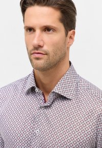 Camicia in cotone a fantasia con base bianca e design geometrici rossi e blu, colletto aperto e bottoni bianchi.