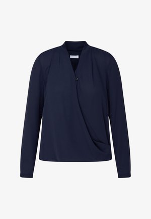 Persoon draagt een marineblauwe blouse met lange mouwen, V-hals en overslagdetail, gecombineerd met een zwarte broek, handen in de zakken.