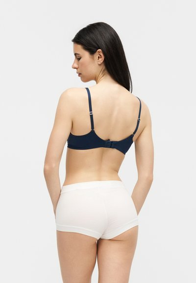 Γυναίκα με μακριά σκούρα μαλλιά που κοιτάζει πλάγια, φοράει μπλε σουτιέν και λευκά εσώρουχα, μπροστά σε απλό λευκό φόντο.