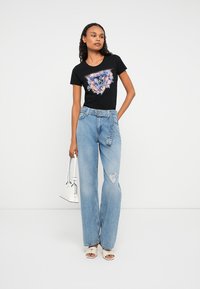 Kvinne står, iført en svart blomstrete Guess-logo t-skjorte, lyseblå høytlivs ripped jeans med belte, hvit håndveske og hvite hæl sandaler.