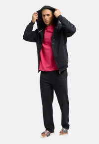 Sort zip-up hoodie med broderede detaljer, pink crew neck t-shirt under, sorte joggers og mønstrede sandaler på en model.