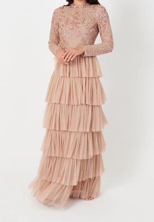 Kobieta ubrana w długą, warstwową beżową suknię z cekinowym gorsetem na długie rękawy i warstwową tiulową spódnicą, stoi z rękami złożonymi razem.