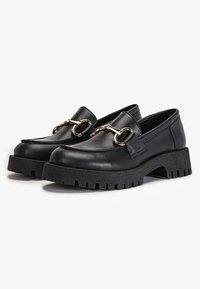 Paire de mocassins en cuir noir avec semelles épaisses à crampons et détail en métal doré en forme de mors de cheval sur le devant.