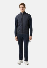 Navy zip-up jacket with stand collar, paired with matching navy pants. Model wears white sneakers and holds sunglasses in hand.