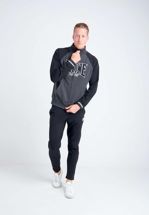 Jeune homme debout portant une veste zippée noire et grise, un pantalon noir et des baskets blanches devant un fond blanc uni.