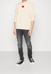 Sudadera beige con cuello redondo y logo cuadrado rojo, combinada con unos vaqueros oscuros y desgastados y zapatillas negras con suelas y cordones blancos.