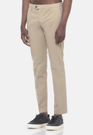 Uomo che indossa pantaloni beige slim fit e sneaker grigie scure, in piedi davanti a uno sfondo semplice.