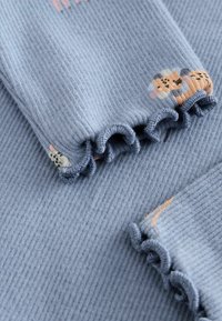 Blaue gerippte Stoff mit kleinen gestickten Tiermotiven und Rüschensäumen. Die Textur ist weich und leicht, ideal für die Freizeit.