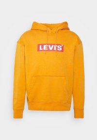 Žlutá mikina s kapucí, klokaní kapsou na přední straně a červeným logem Levi's na bílém pozadí. Měkká textilie.